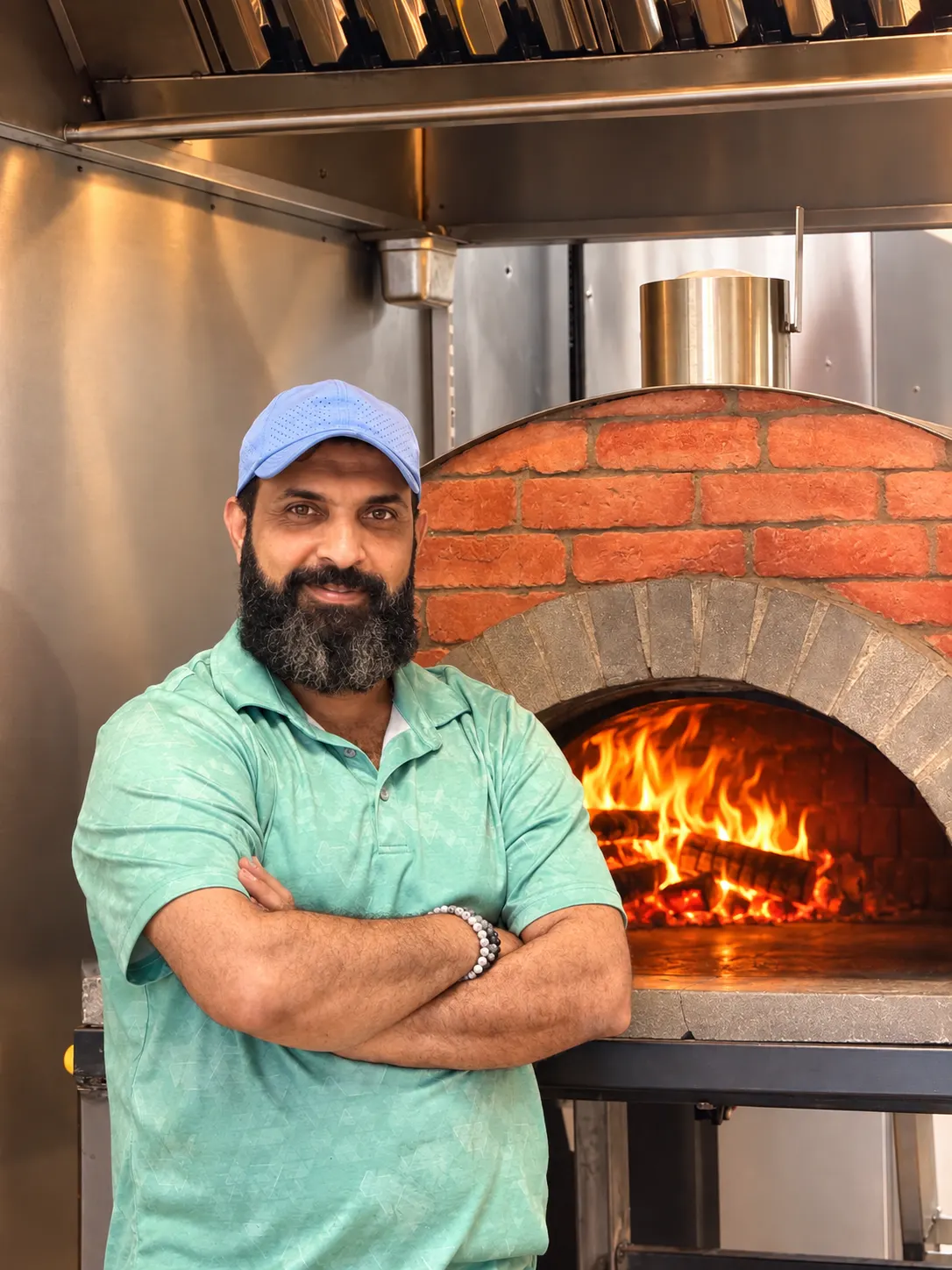 Fouad standing with his arms crossed in front of the brick half-dome oven, fire burning inside