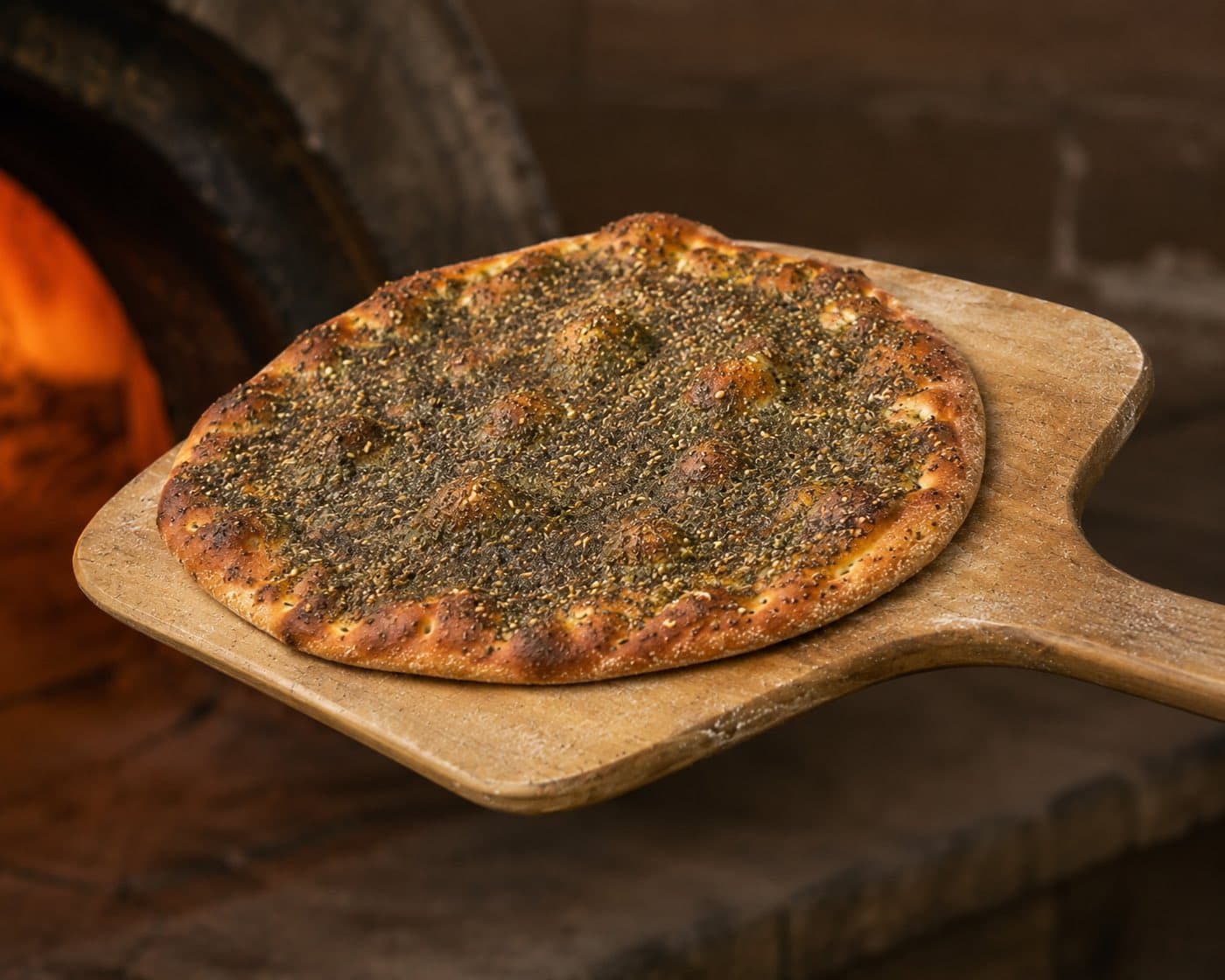 A za'atar manoush on a wooden peel, just pulled from the stone oven with the fire still burning behind it