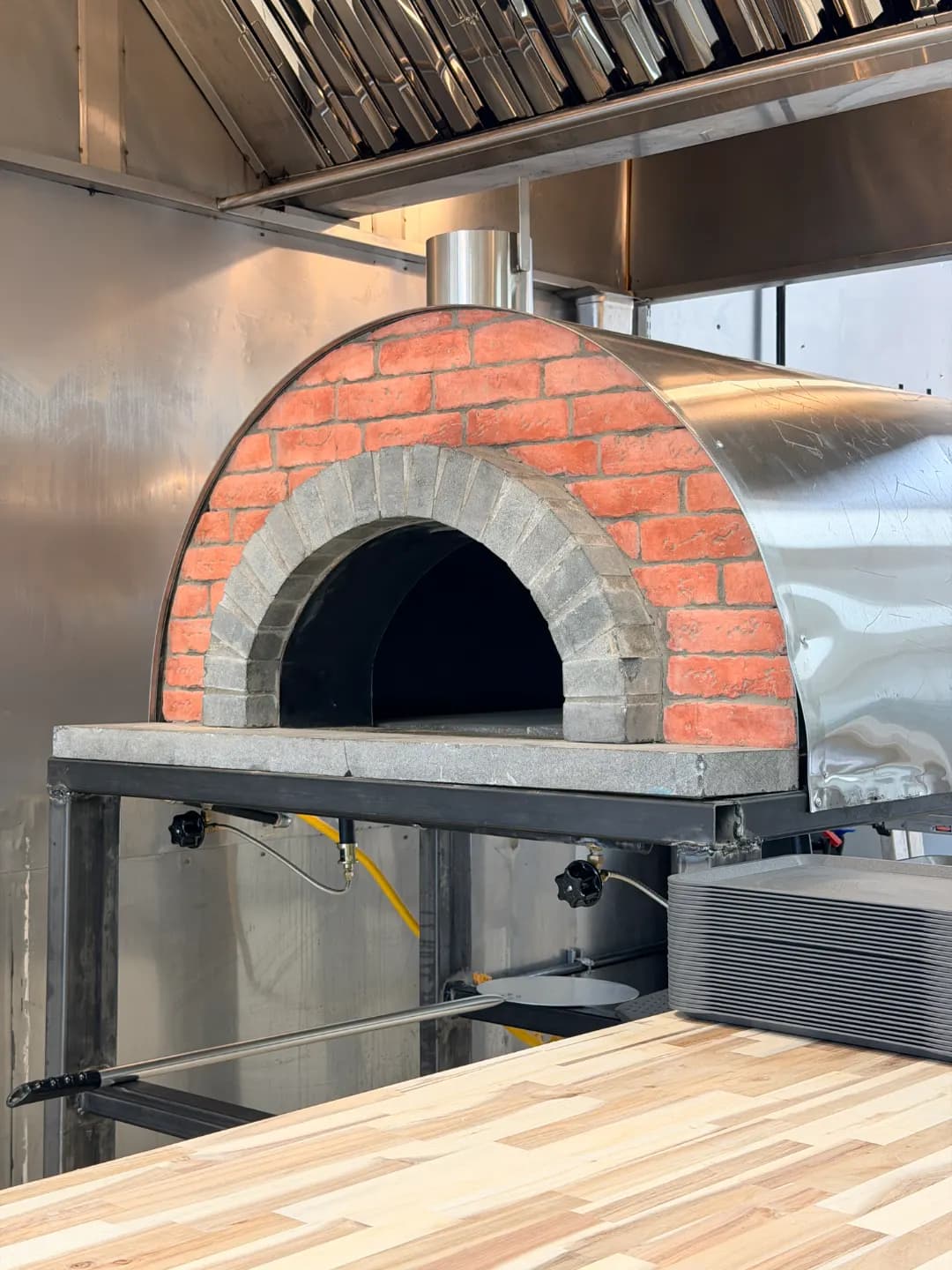 The half-dome oven from the side: red brick face, grey stone arch and steel body on a welded stand, over a butcher-block counter